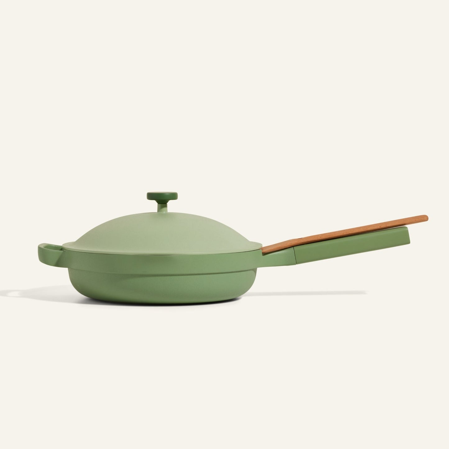 The Always Pan 2.0, a green multi-purpose skillet with a lid and long wooden handle, is displayed on a plain light background, showcasing its versatility as part of a bestselling cookware system.