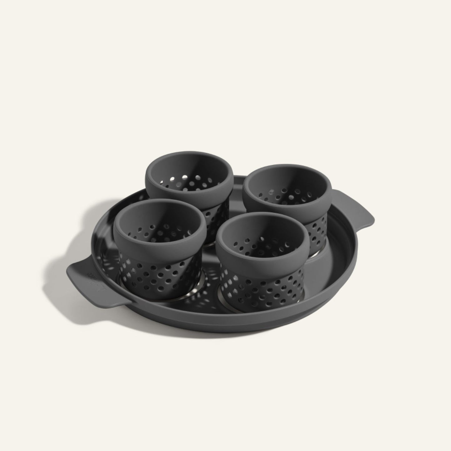 The Egg Poacher features a round tray with handles holding four black silicone cups for poaching eggs—perfectly arranged and an ideal accessory for the Always Pan.