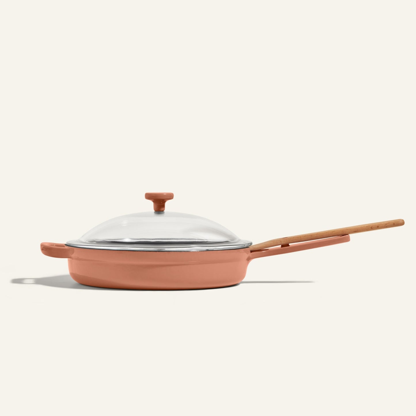 The Cast Iron Always Pan, a pink enameled nonstick skillet with a glass lid and wooden handle, offers 8-in-1 versatility and is displayed against a plain off-white background.
