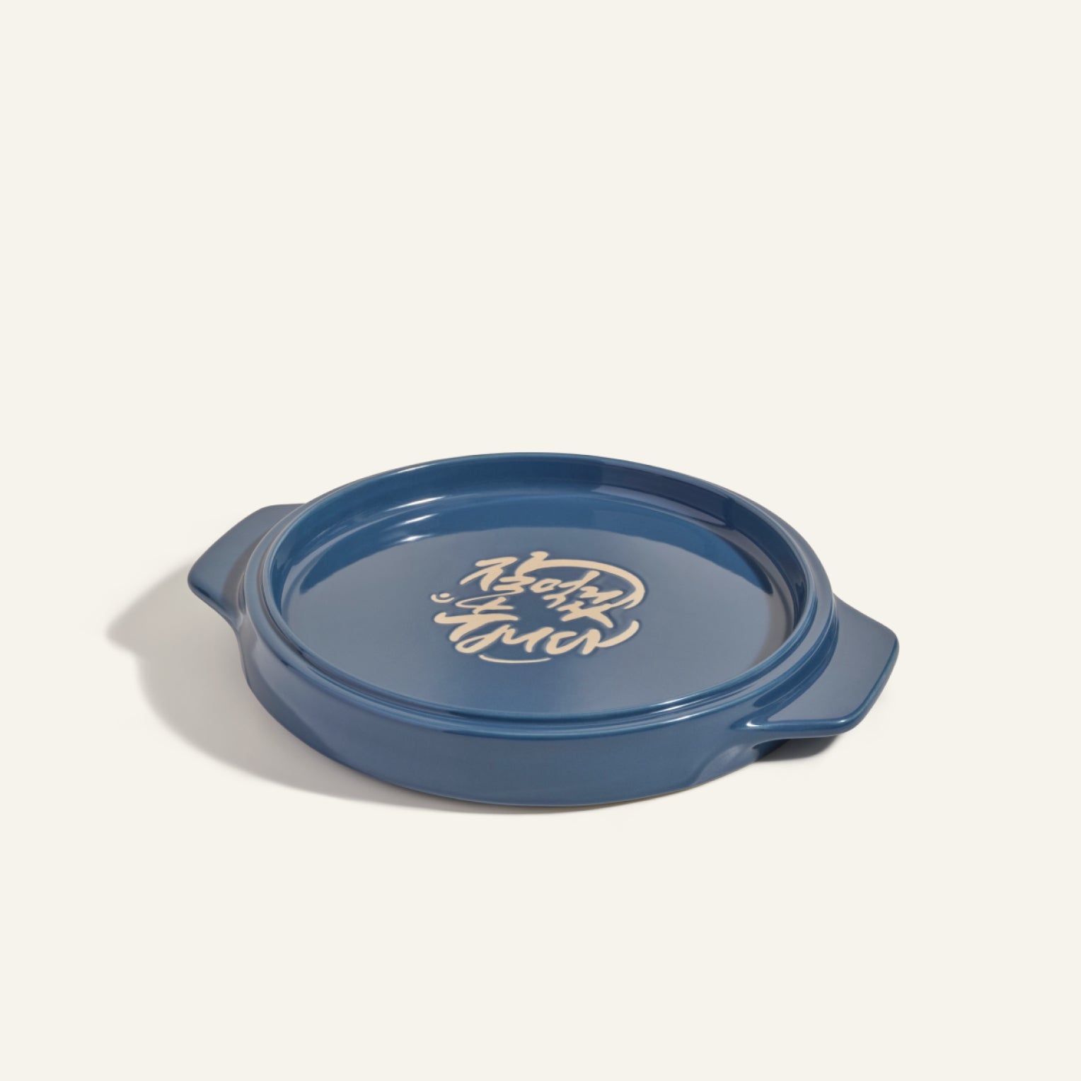 The Flipping Platter is a round blue ceramic dish with two side handles and white Korean writing on the base, ideal for serving maqluba or tahdig, shown against a plain background.