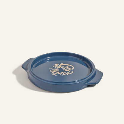 The Flipping Platter is a round blue ceramic dish with two side handles and white Korean writing on the base, ideal for serving maqluba or tahdig, shown against a plain background.
