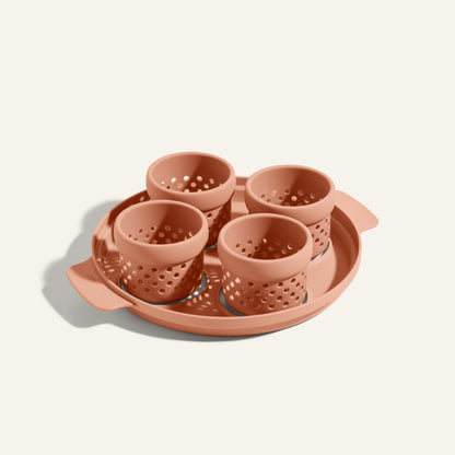A set of four terracotta-colored Egg Poacher silicone cups arranged on a matching round tray with handles, against an off-white background.