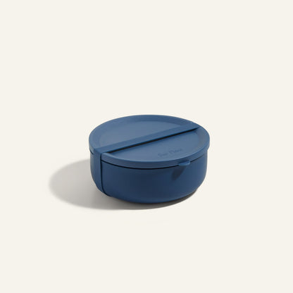 The Wander Bowl is a round, blue plastic lunch container with a fitted lid, shown against a plain background. Microwave safe, it's ideal for storing and reheating meals.