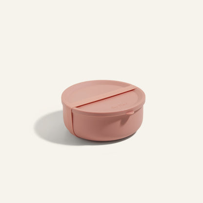 The Wander Bowl, a round pink lunch container with a matching lid, is shown on a plain background. This microwave-safe solution keeps your meals fresh and ready to enjoy.