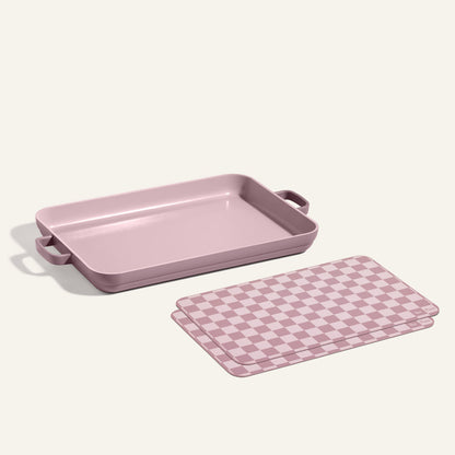 A Griddle Pan with handles sits next to Oven Mats, both featuring mauve and white checkered patterns, on a neutral background.