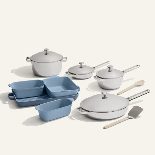 The VIP Titanium Pro Complete Cookware + Bakeware Set features stainless steel pots, titanium pans with lids, nonstick blue baking dishes and loaf pans, plus a wooden spoon and metal spatula, all arranged on a light surface.