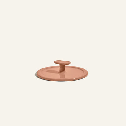 A terracotta-colored, round ceramic lid with a central handle—resembling the Grill Press—is displayed against a plain light background, highlighting its even heat distribution.